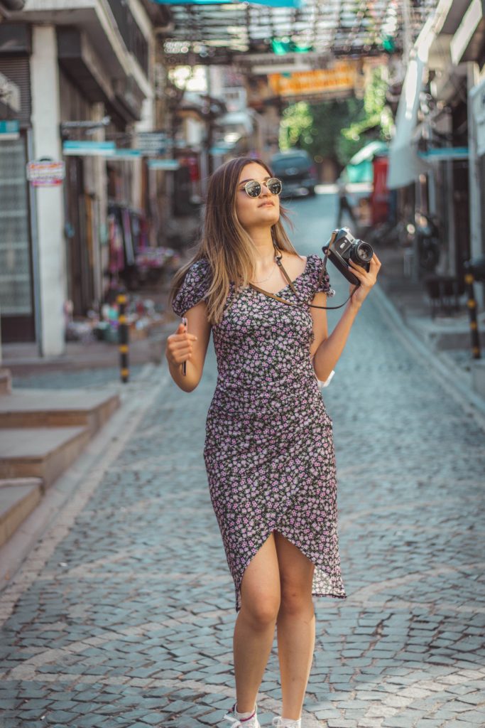 Stylish woman in floral dress exploring Beypazarı street with camera, capturing lifestyle moments.